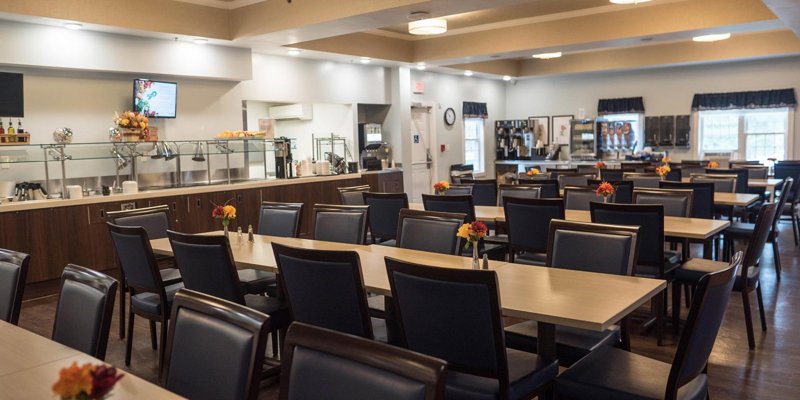 A spacious and welcoming dining area with neatly arranged tables and chairs, warm lighting, and a buffet-style setup, creating a comfortable environment for meals.