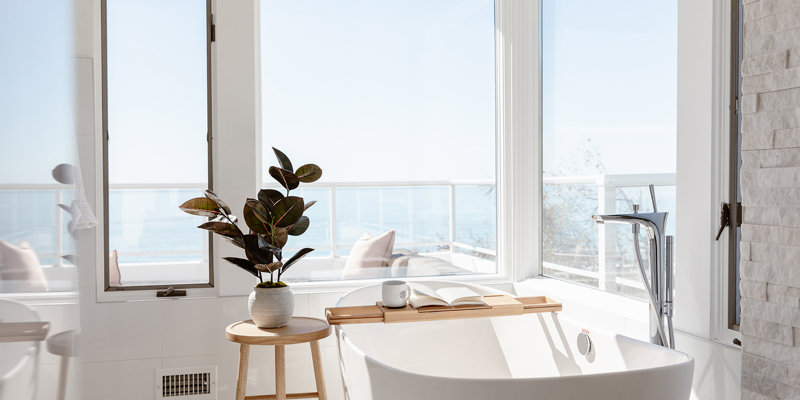 A serene bathroom with a freestanding tub, natural light, and ocean views, complemented by a plant on a wooden stool for a relaxing atmosphere.