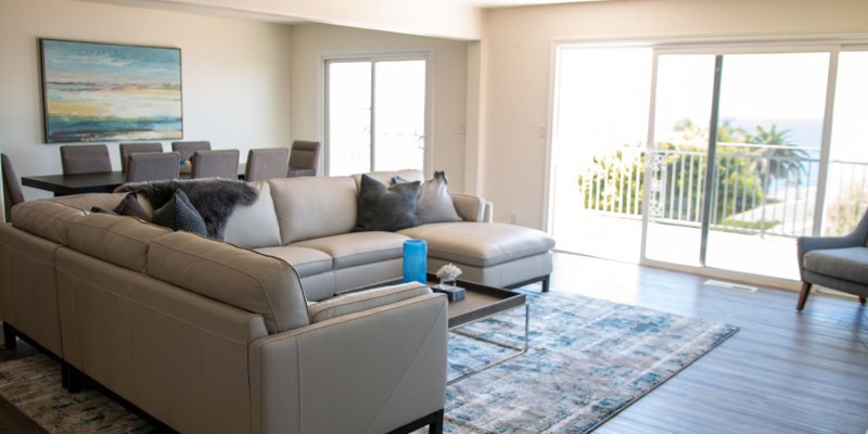 Bright and open living space featuring a modern sectional sofa, large dining table, and expansive ocean view through sliding glass doors.