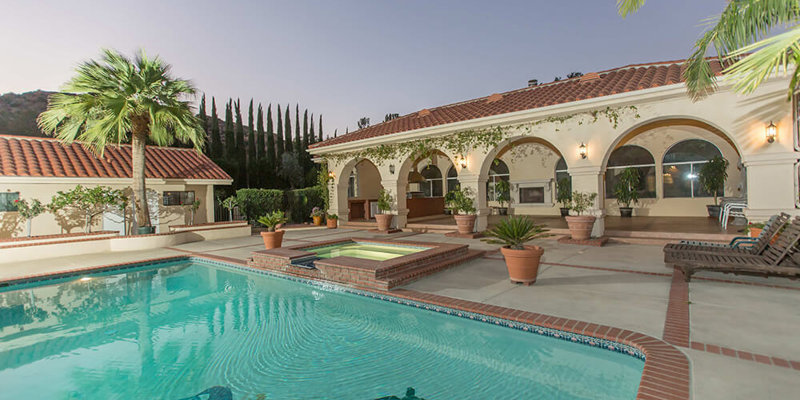 A serene backyard oasis with a spacious pool, hot tub, and lush greenery, surrounded by elegant arches, palm trees, and cozy lounging areas.