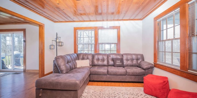 A cozy living room with a plush gray sectional sofa, warm wooden ceilings, large windows, and red ottomans, creating a comfortable and inviting space.