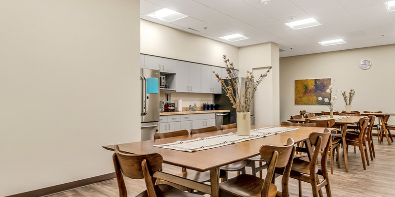 A spacious dining area featuring light wood tables and chairs, with soft lighting and a modern kitchen in the background, offering a welcoming environment.