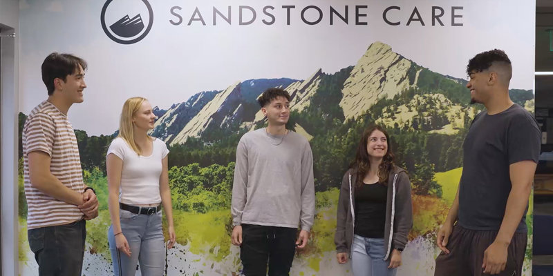 A group of four people smiling and standing together in front of a vibrant Sandstone Care backdrop featuring scenic mountain views.