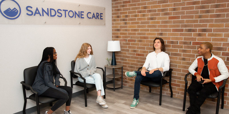 A group of four young adults sit comfortably in a modern waiting area at Sandstone Care, engaging in casual conversation by a brick wall.