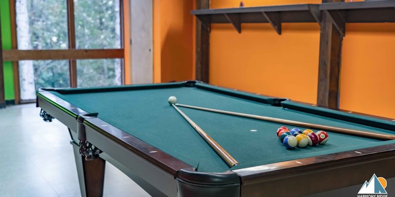 Inviting game room featuring a pool table with cues and balls ready for play, set against a warm, orange accent wall and large windows.