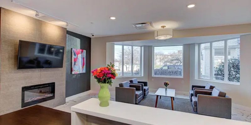 A cozy, well-lit lounge area featuring comfortable seating, a flat-screen TV, a modern fireplace, and a vase of fresh flowers on the counter.