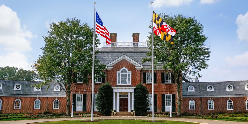 A grand brick building with colonial architecture, flanked by trees, and proudly displaying the American and state flags on a sunny day.