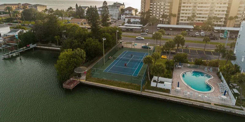 An aerial view of a scenic waterfront property featuring a tennis court, a kidney-shaped pool, and lush greenery, with nearby buildings and roads.