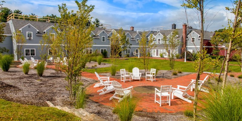 A beautiful outdoor space with Adirondack chairs arranged around a fire pit, surrounded by greenery, offering a serene environment for relaxation and socializing.