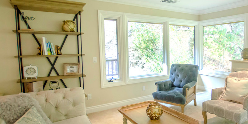 A bright and cozy living room featuring plush seating, a wooden coffee table, decorative shelving, and large windows offering serene nature views.