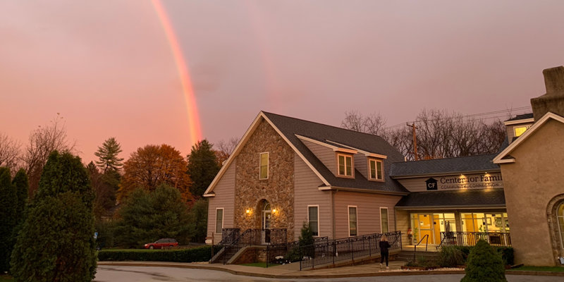 A cozy building with a stone facade sits under a vibrant double rainbow at sunset, surrounded by trees and greenery, creating a serene atmosphere.