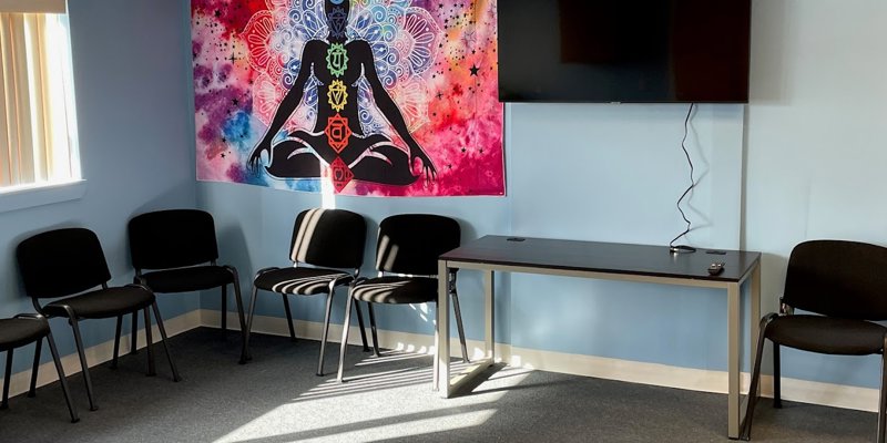 A peaceful room with chairs arranged in a circle, a vibrant tapestry featuring chakra symbols, and a wall-mounted TV, perfect for group sessions.