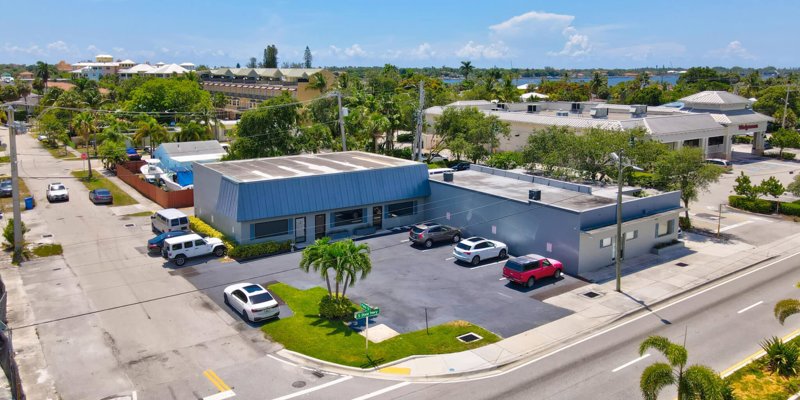 A bright day in a calm neighborhood, with a building and parking lot surrounded by palm trees, close to water and other businesses.