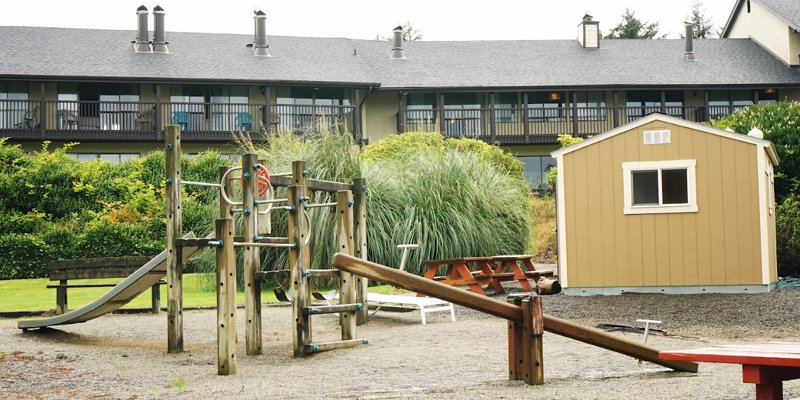 A charming outdoor play area with a wooden playground, slide, picnic tables, and a storage shed, surrounded by greenery and a cozy resort.