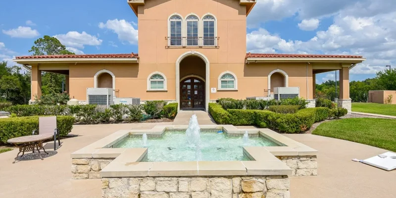 A beautiful Mediterranean-style building with arched windows and a charming fountain in front, surrounded by lush greenery and a relaxing outdoor area.