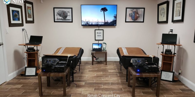  A specialized therapy room featuring two treatment beds, advanced brain stimulation equipment, framed artwork, and a calming wall-mounted TV display.