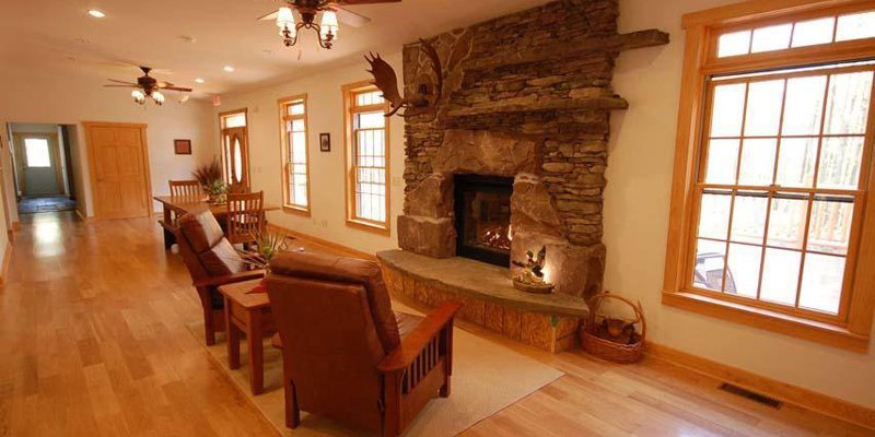 A warm, inviting room with a rustic stone fireplace, wooden floors, and large windows offering natural light, perfect for relaxation.