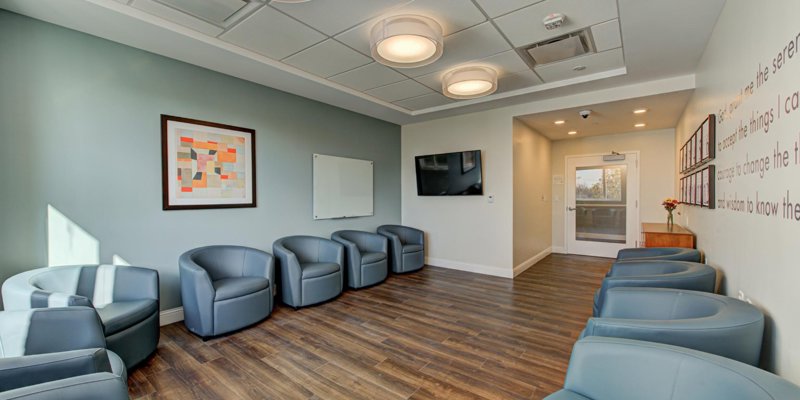 A cozy, well-lit meeting room with comfortable blue chairs, wood flooring, modern artwork, a TV, and inspiring wall quotes for a peaceful atmosphere.