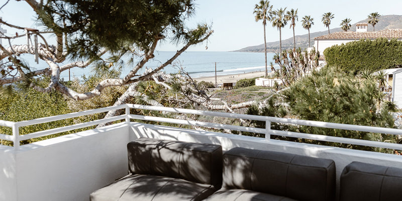 Cozy outdoor seating area with a modern sofa, wooden table, and stunning ocean views framed by palm trees and lush greenery.