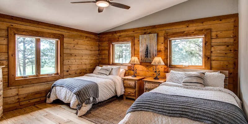 A cozy cabin bedroom with two comfortable beds, wooden walls, large windows offering scenic views, and soft lighting, creating a peaceful retreat.