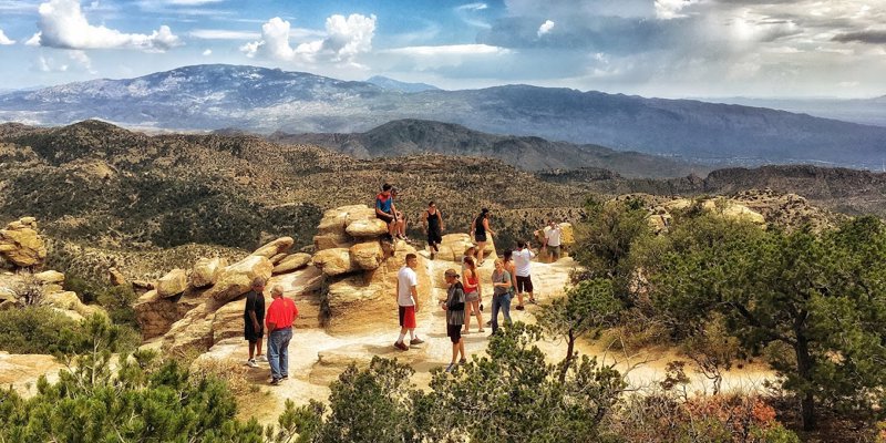 A group of people enjoy a scenic hike atop rocky cliffs with expansive views of rolling mountains, under a sky with dramatic clouds.