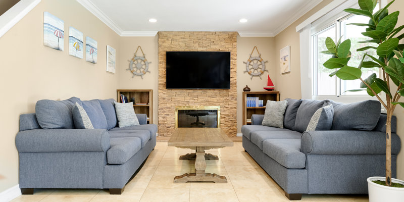 A cozy living room with two plush gray sofas, a wooden coffee table, and a stone fireplace with a mounted TV, decorated in a nautical theme for relaxation.