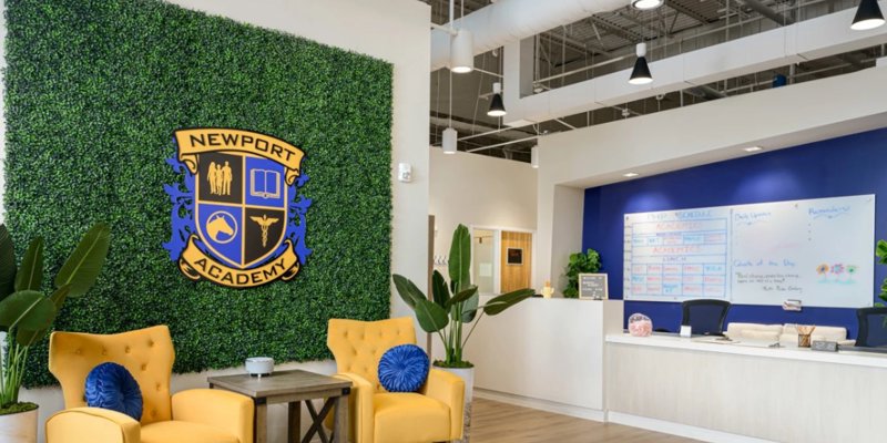 A bright and welcoming reception area features a Newport Academy crest, vibrant yellow chairs, greenery, and a modern, organized desk space.