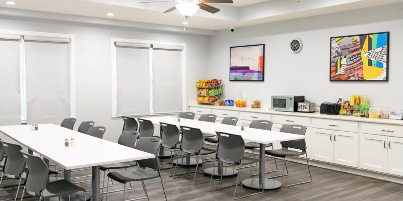 Bright dining area with modern decor, long tables, and plenty of seating, featuring a snack station and colorful artwork on the walls.