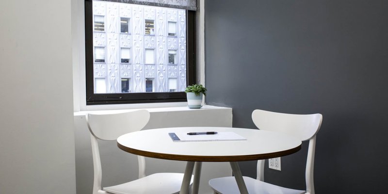 A small, modern meeting area with a round white table, two chairs, and a large window, creating a simple and focused space for discussions.