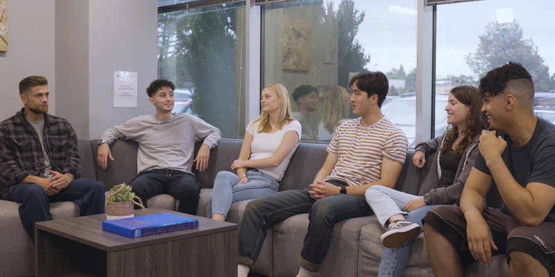 A group of six people sits comfortably on a gray sofa in a bright room with large windows. They appear relaxed, engaged in a friendly discussion.