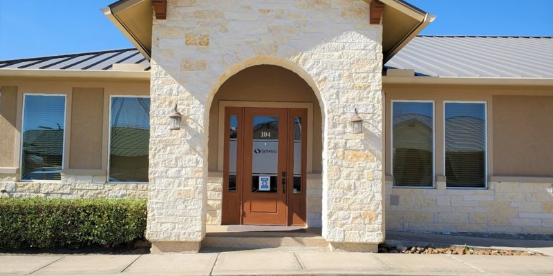 Welcoming entrance of a clinic with a stone archway, warm wooden doors, and well-maintained landscaping under a clear blue sky