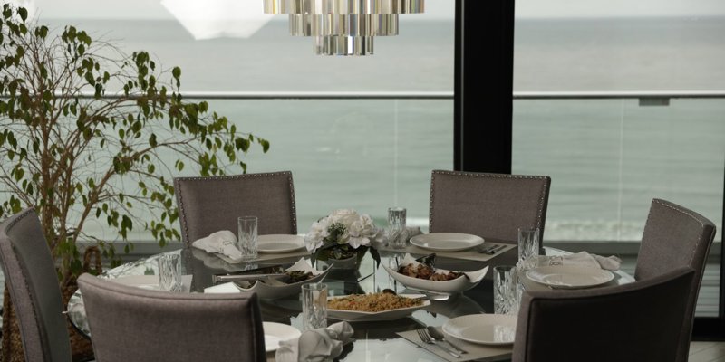 Elegant dining setup with a glass table, plush chairs, and ocean views. A crystal chandelier adds sophistication to the serene ambiance.