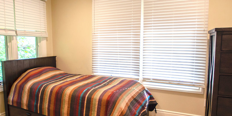 A cozy bedroom featuring a single bed with a colorful striped quilt, large windows with blinds, and a warm, inviting atmosphere for rest and relaxation.