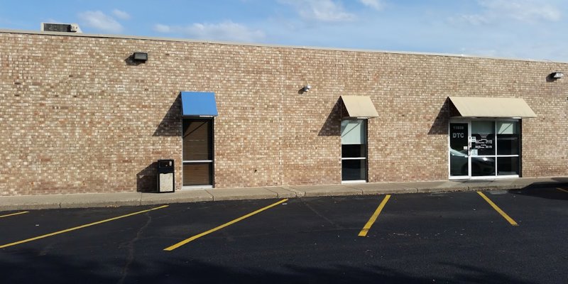 Brick building with large windows and awnings, featuring a welcoming entrance and ample parking space under a clear, blue sky.