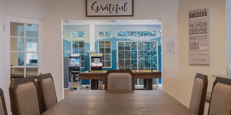 A welcoming dining area with a "Grateful" sign above the entrance to a game room, featuring natural light, arcade games, and bright decor.