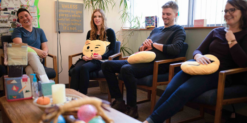 A cheerful group sits in a cozy room, holding emoji pillows, engaging in a supportive, light-hearted discussion surrounded by uplifting decor.