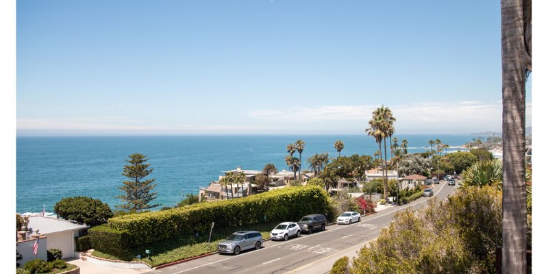 Scenic coastal view featuring palm trees, a blue ocean, and a sunlit road with cars driving along, highlighting a peaceful seaside atmosphere.