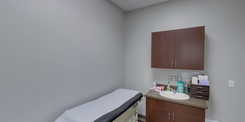 A simple, clean medical exam room with an examination table, wooden cabinets, a sink, and essential supplies neatly arranged for patient care.