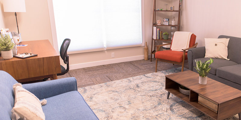 A cozy and inviting office lounge with a blue sofa, a gray loveseat, a wooden desk, a bookshelf, and soft, natural lighting creating a calm ambiance.