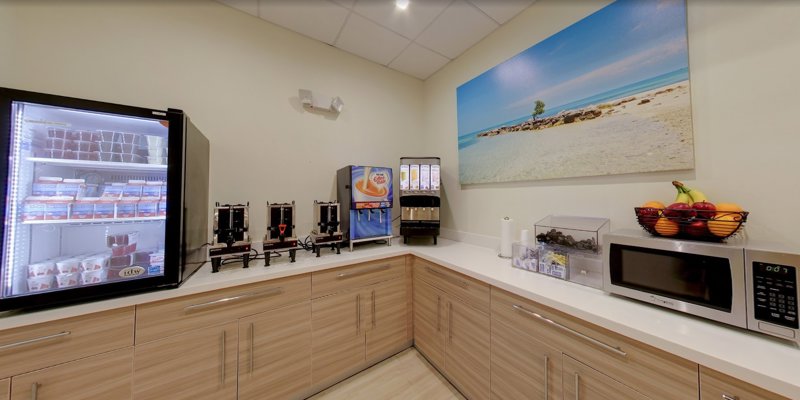 A bright kitchenette featuring a fridge with assorted drinks, coffee dispensers, and a microwave. A vibrant beach photo enhances the space.