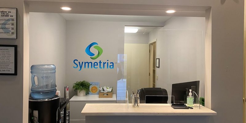 The reception area is clean and inviting, featuring a water cooler and modern decor. The Symetria logo adds a professional and welcoming touch.