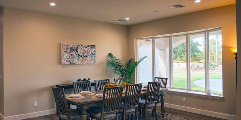 Elegant dining room with a large wooden table set for eight, natural light streaming through a wide window, and tasteful décor including a potted plant and artwork.