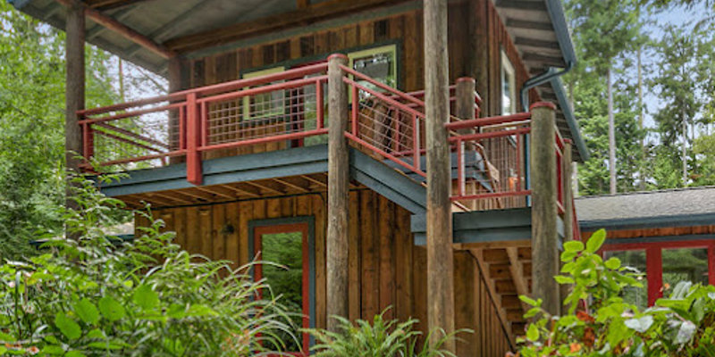 A serene, rustic cabin surrounded by lush greenery, featuring a wooden balcony and stone pathway, offering a peaceful and inviting retreat.