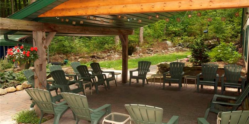  A serene outdoor gathering space with green Adirondack chairs arranged under a rustic wooden pavilion, surrounded by lush greenery.