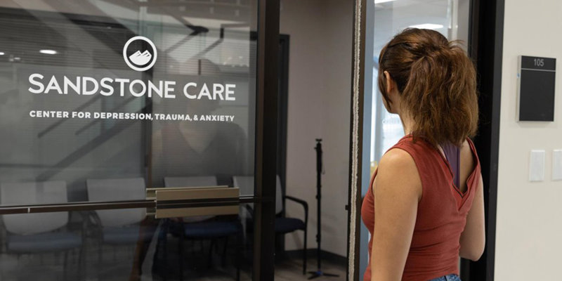  A person stands outside the entrance of Sandstone Care, a center for depression, trauma, and anxiety, creating a welcoming, supportive space.